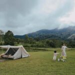 Une tente dressée dans un pré avec deux enfants jouant près d'une rivière et montagnes en arrière-plan